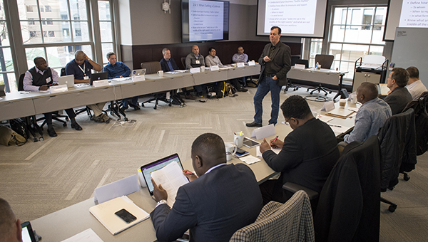 A classroom of executives listens to a teacher standing in the middle of the room