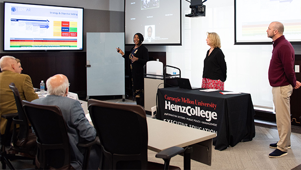 Three people present to an executive education class. The speaker points at the Powerpoint image on the screen.