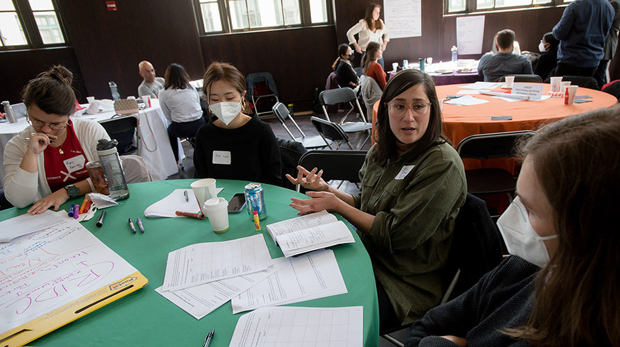 Four people sit around a table covered in papers and discuss their project